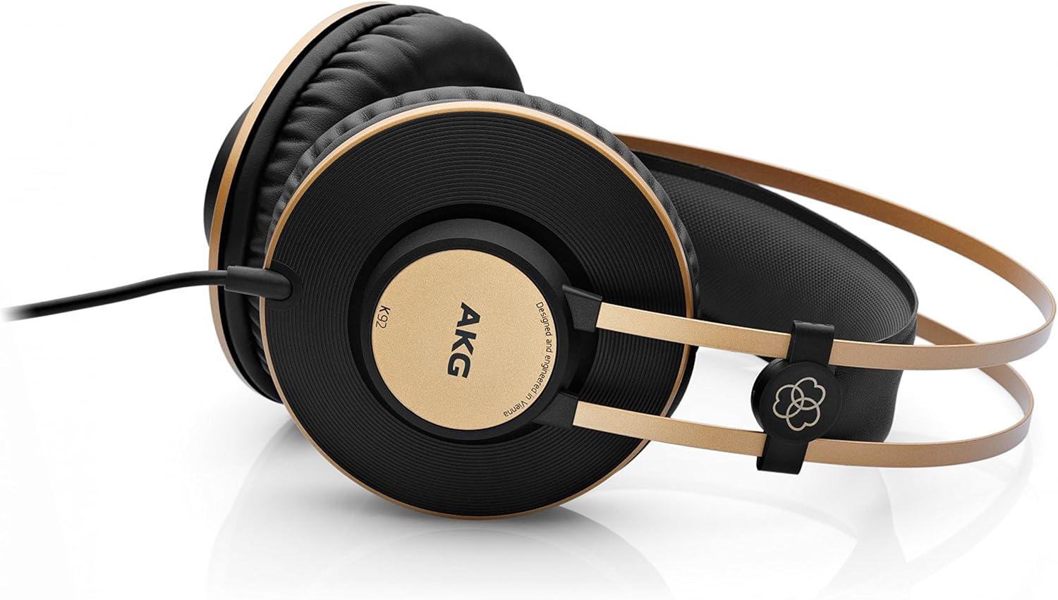 Fone AKG K92 deitado com almofadas grandes pretas, anéis dourados nas laterais, arco duplo dourado flexível e cabo preto, refletido em superfície branca.