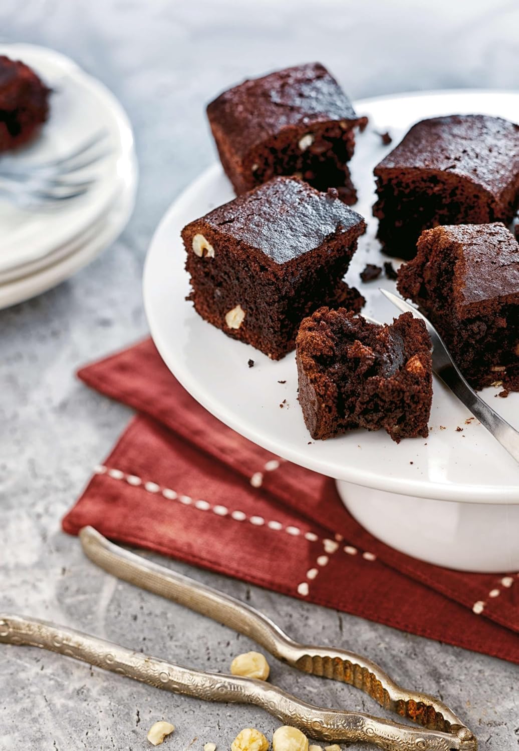 Several square-shaped chocolate brownies with nuts, some on a white cake stand and one with a bite taken out, demonstrating baked goods made with the insert.