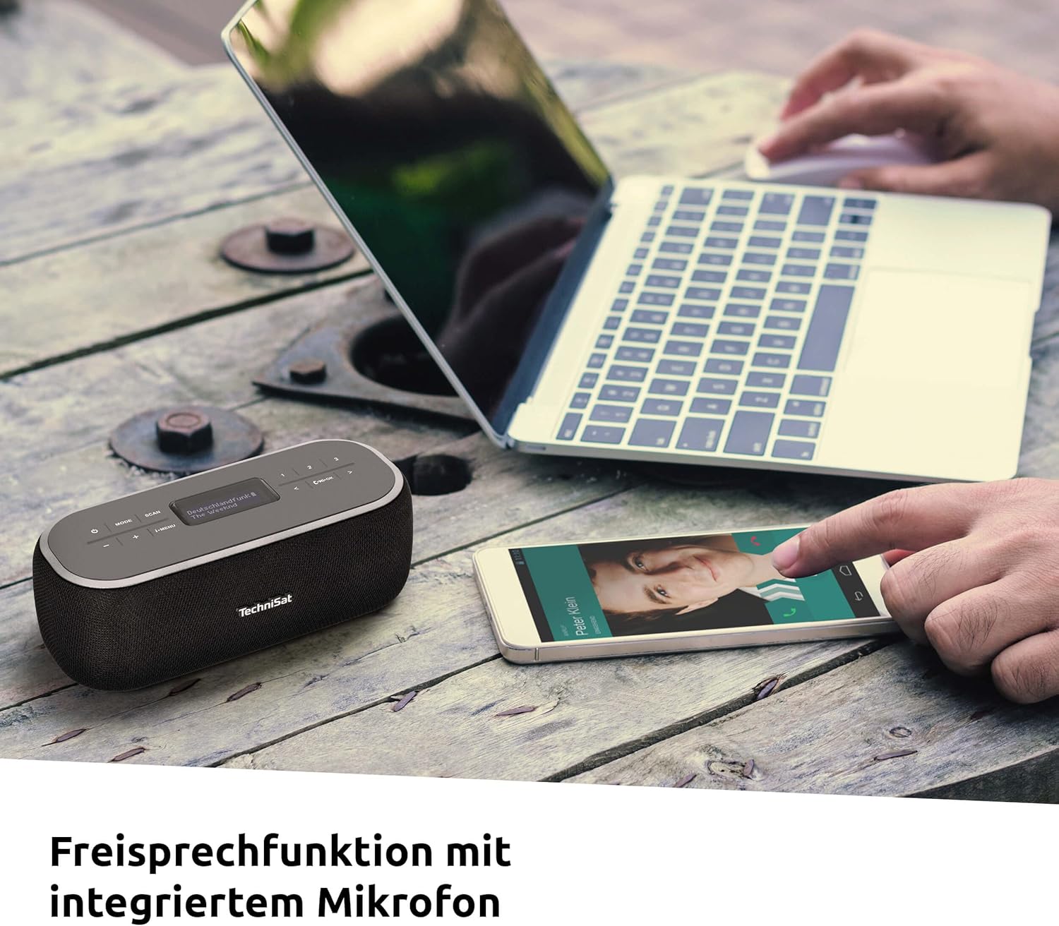 TechniSat VIOLA BT 1 on a wooden table with a laptop and a smartphone displaying an incoming call, indicating the hands-free function.