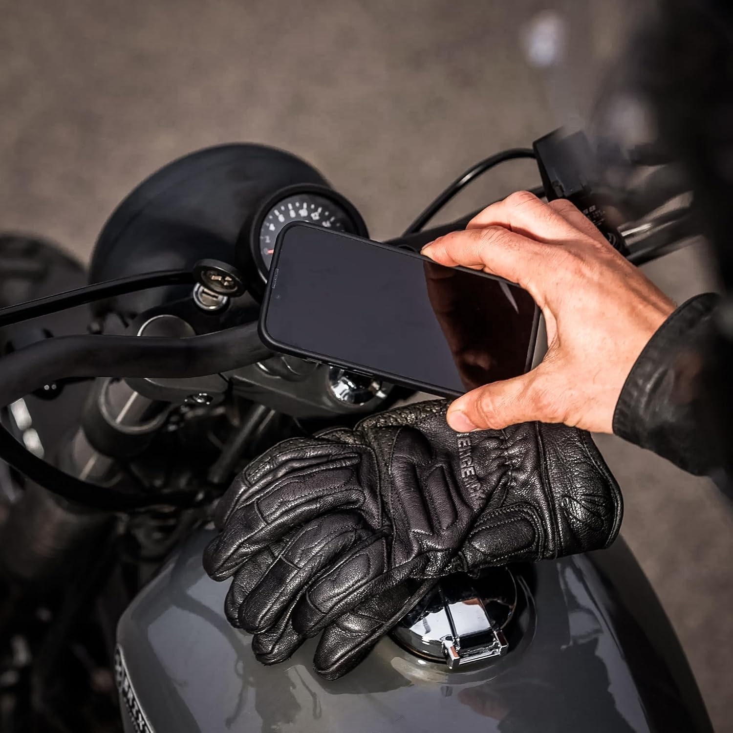 A hand detaching a smartphone from the SP Moto Mount LT on a motorcycle, with a leather glove visible.