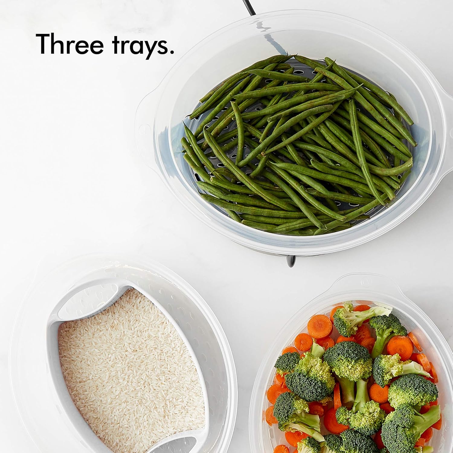 Three clear plastic steaming trays, one filled with green beans, another with broccoli and carrots, and a rice bowl with uncooked rice.
