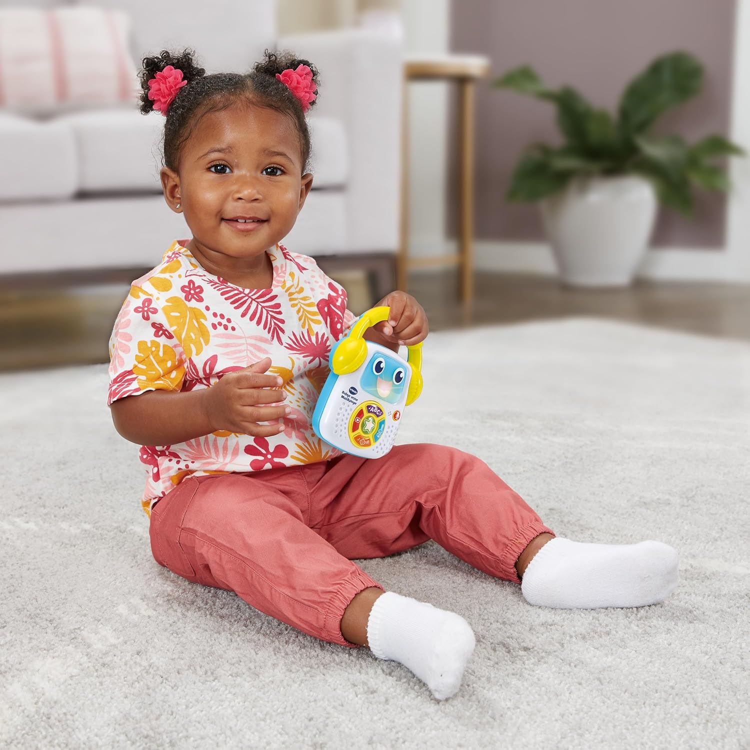 Toddler girl sitting on a rug, happily holding and interacting with the VTech Baby's First Music Player.