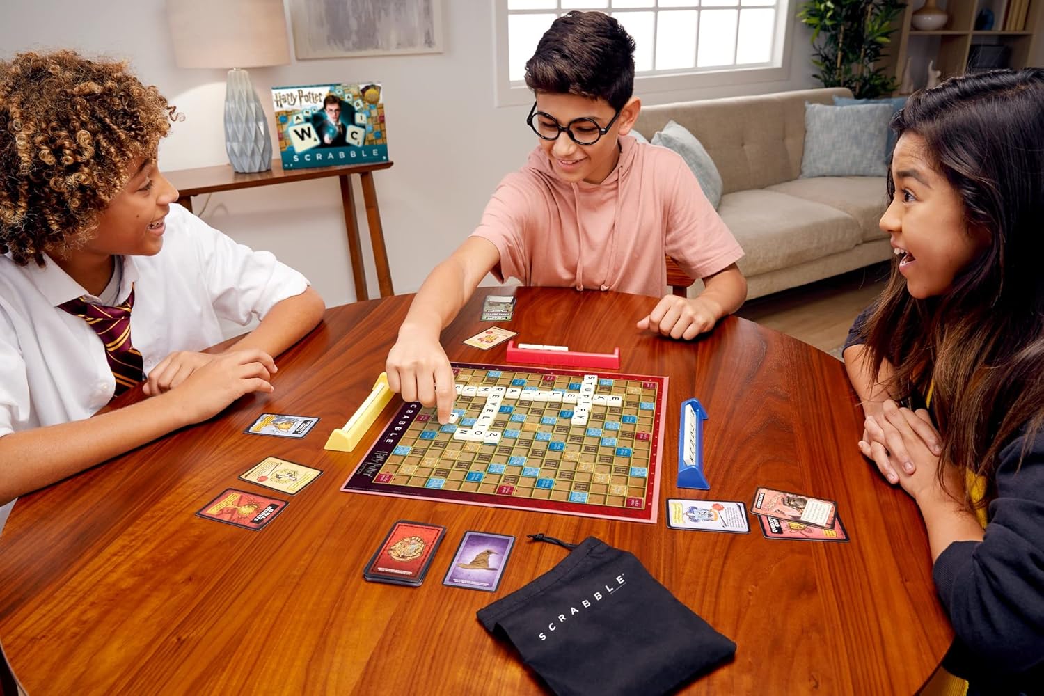 Three children playing the Scrabble Harry Potter game around a wooden table, with the game board, tiles, and cards visible