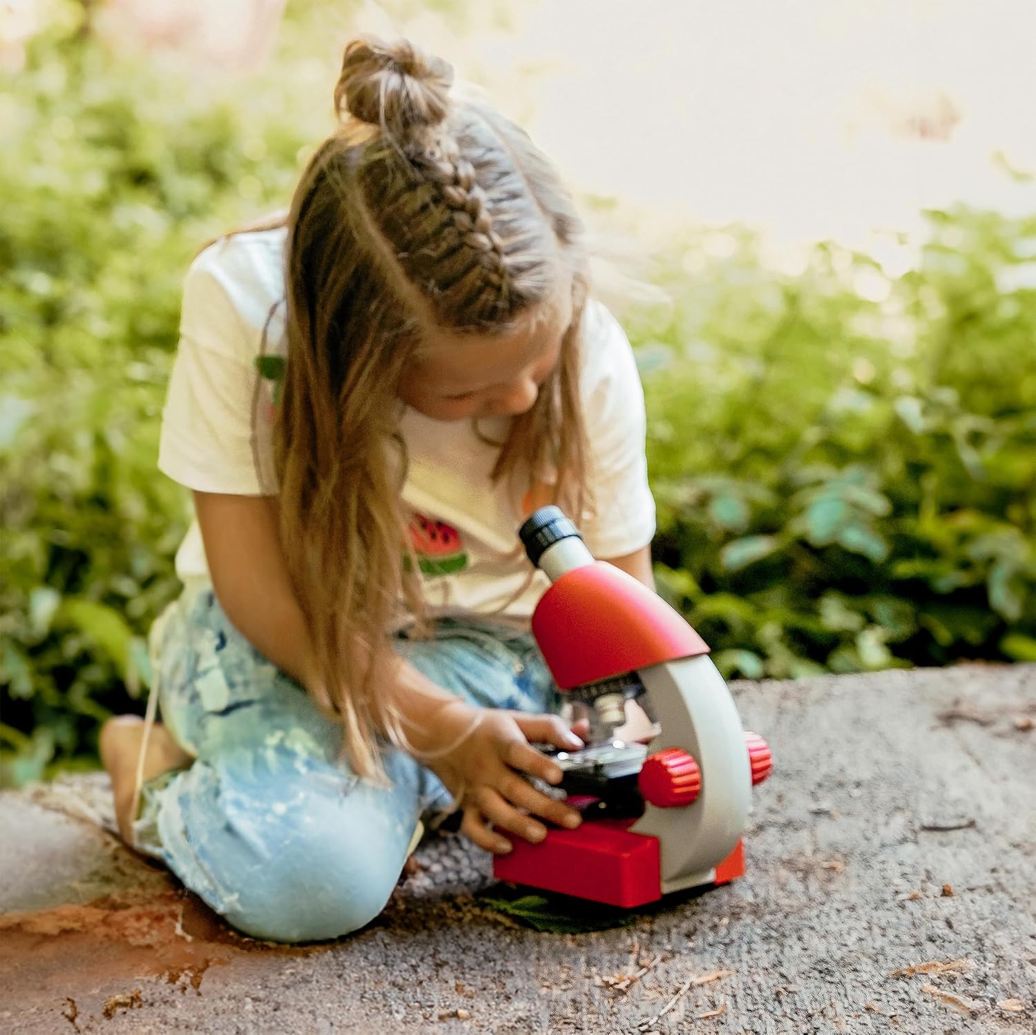 A child with braided hair intently looking through the eyepiece of a red Bresser Junior Microscope, demonstrating typical user interaction.