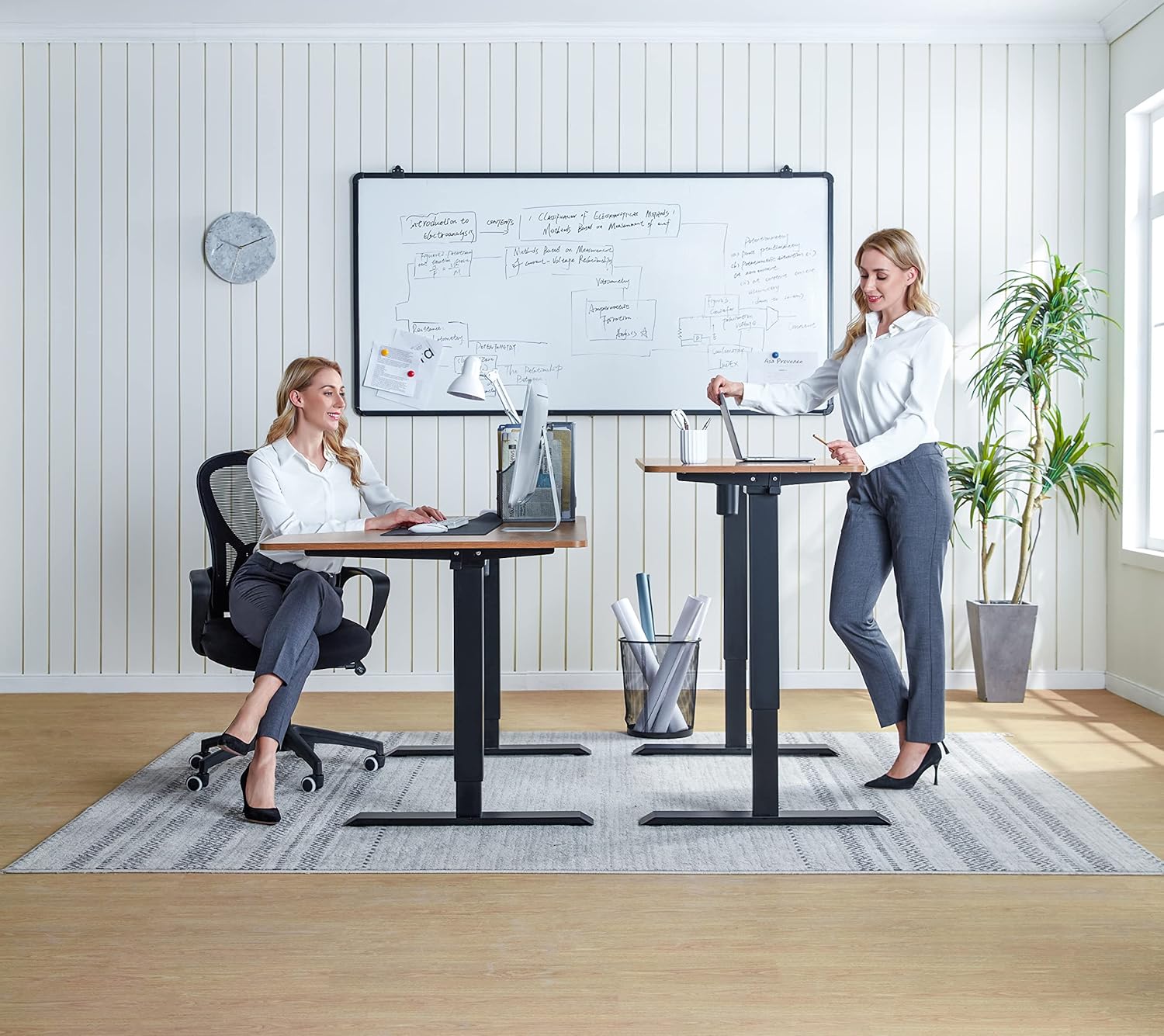 Two women working at LINSY L Shaped Electric Standing Desks, one sitting and one standing, showcasing the height adjustability.