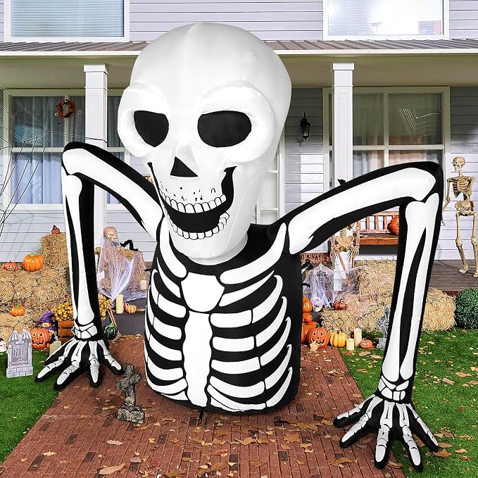Large inflatable skeleton Halloween decoration displayed on a brick front walkway. The white and black skeleton appears to be rising from the ground, with arms spread wide and a grinning skull, surrounded by hay bales, pumpkins, and other Halloween decor.