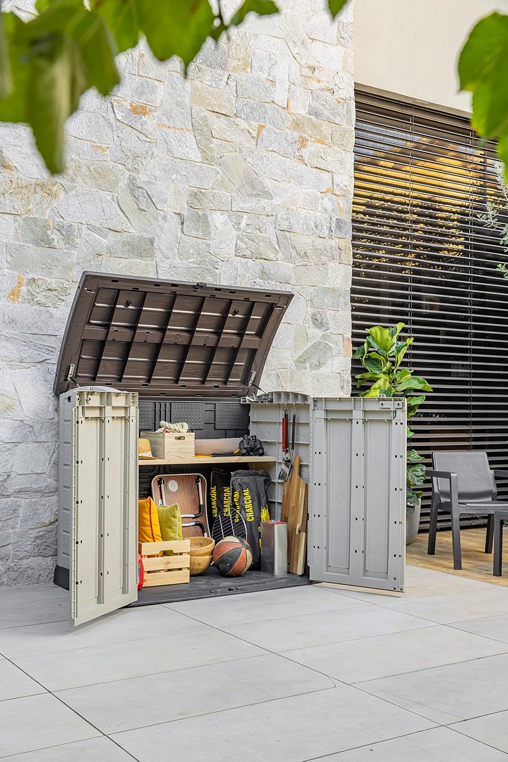 Keter 12-922631 outdoor storage box with lid and doors open, revealing various garden tools and items stored inside.