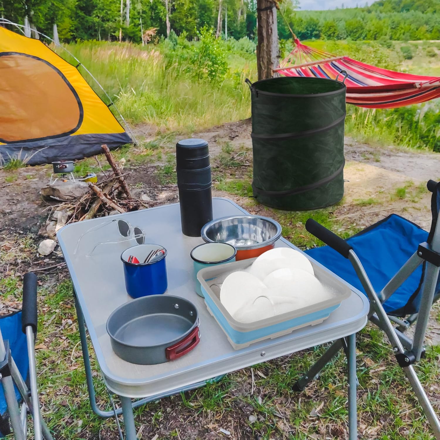 Camping scene with a table, chairs, the collapsible camp sink, and the trash can in the background.