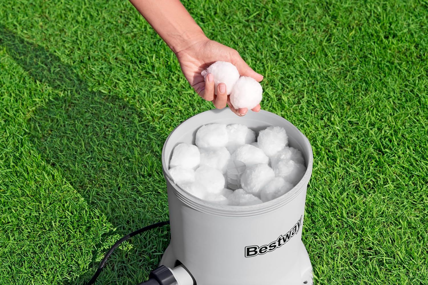 A hand dropping white Polysphere filter balls into the open top of the Bestway filter pump, which is grey with a transparent lid.