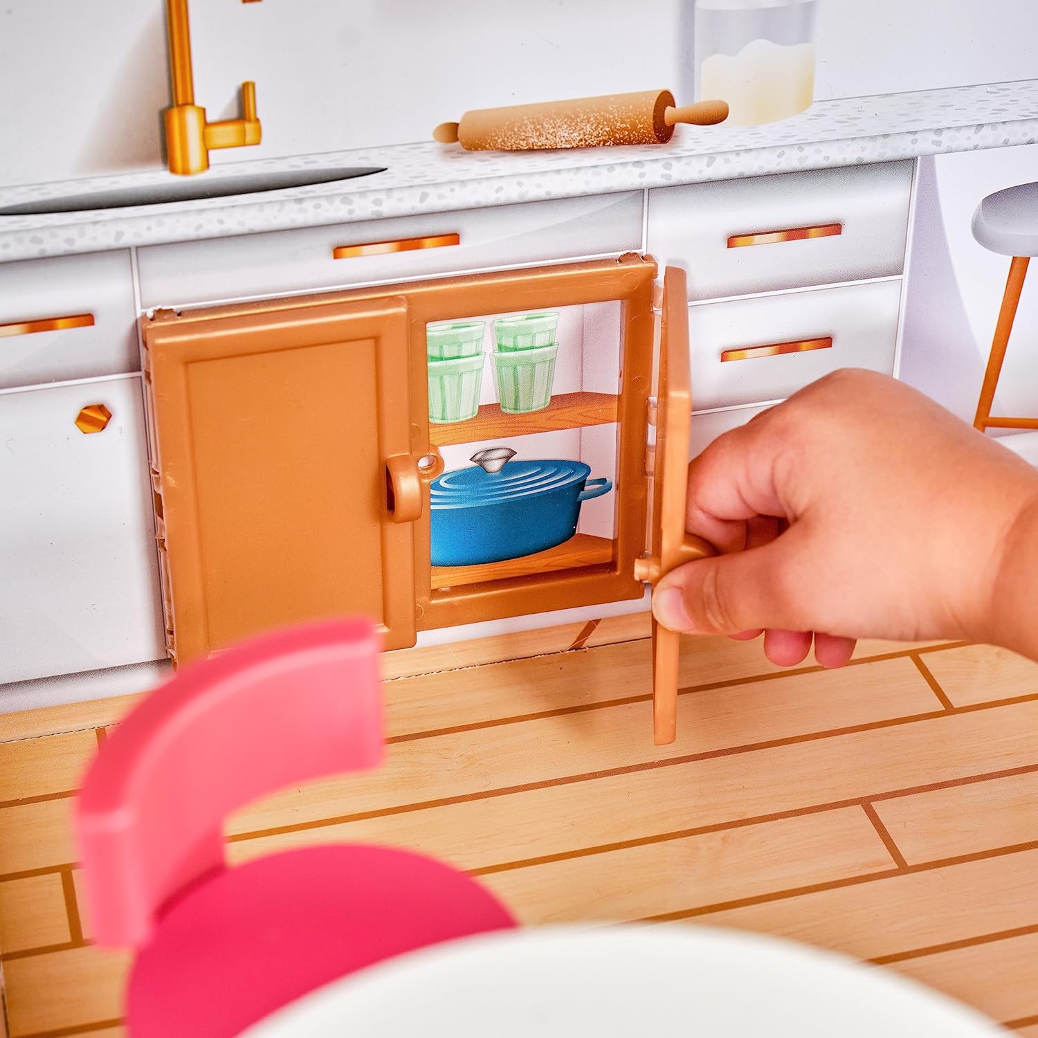 A hand opening a small brown cabinet door in the kitchen area of the dollhouse, revealing miniature dishes inside.