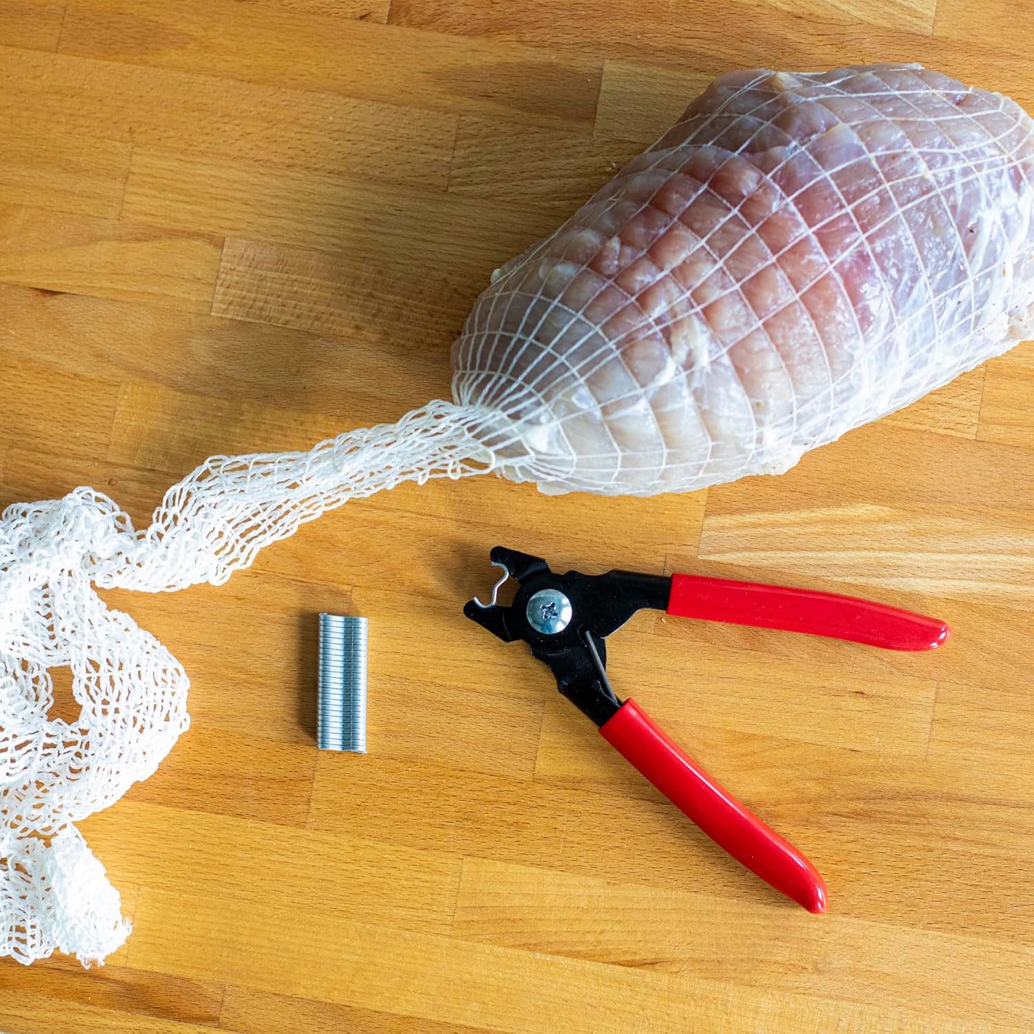 Browin 313313 Sausage Clipper with a stack of metal clips and a netted piece of meat on a wooden surface.