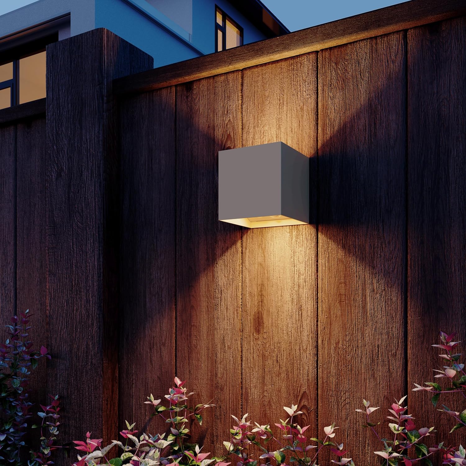 A Calex Volterra cube wall light mounted on a wooden fence at night, casting warm light upwards and downwards, illuminating the surrounding area and plants.