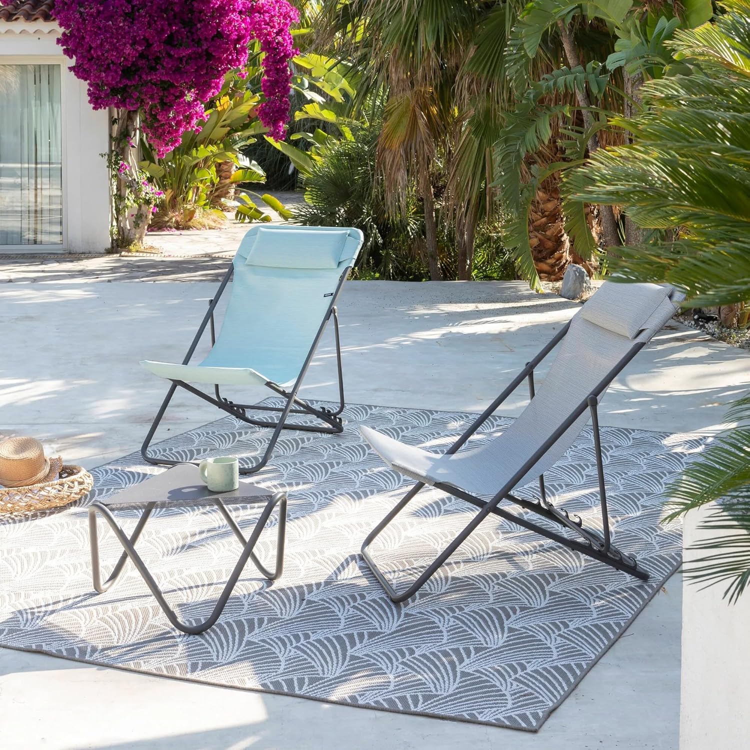 Two Lafuma chaise longues, one grey and one light blue, set up outdoors on a patio with lush greenery.