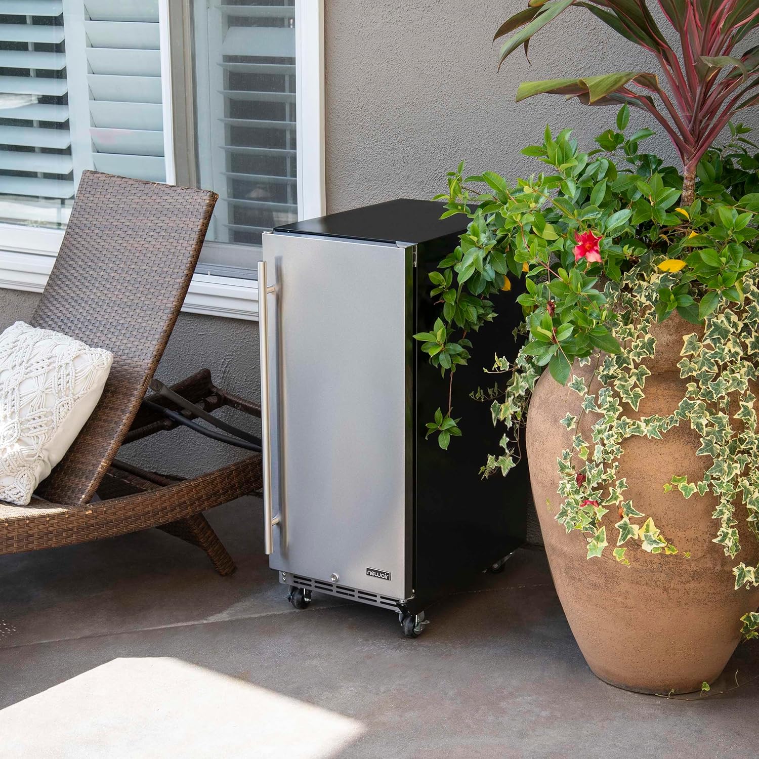 NewAir outdoor beverage refrigerator placed freestanding on a patio next to a lounge chair and a large potted plant.