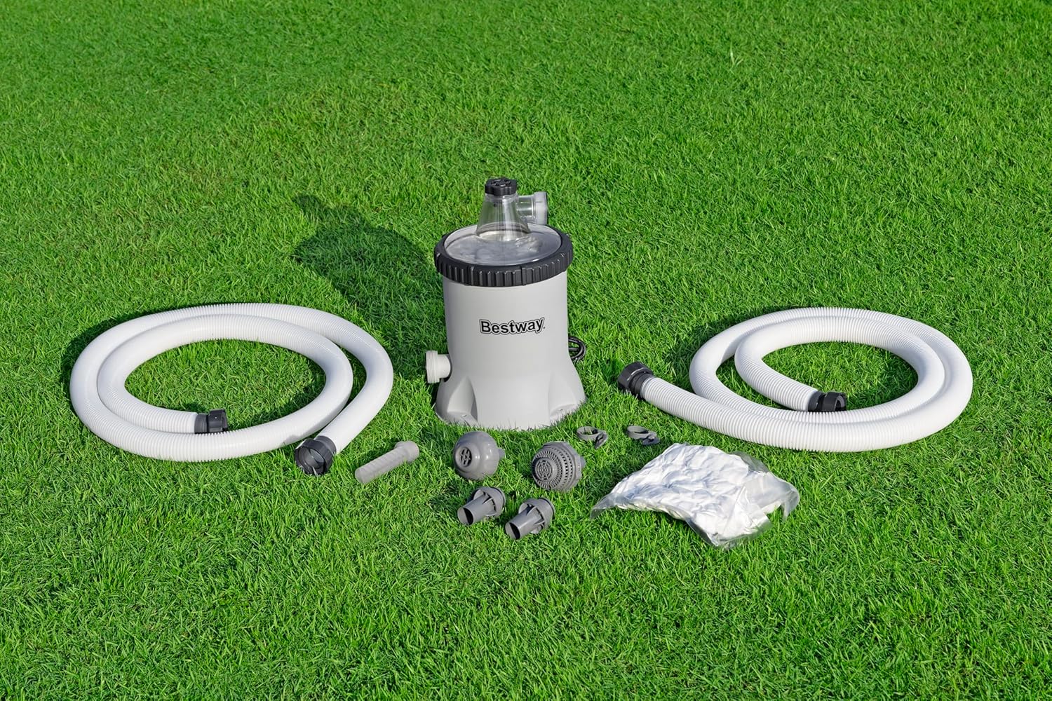 All components of the Bestway filter pump laid out on green grass, including the pump unit, two white hoses, various adapters, and a bag of white Polysphere filter balls.