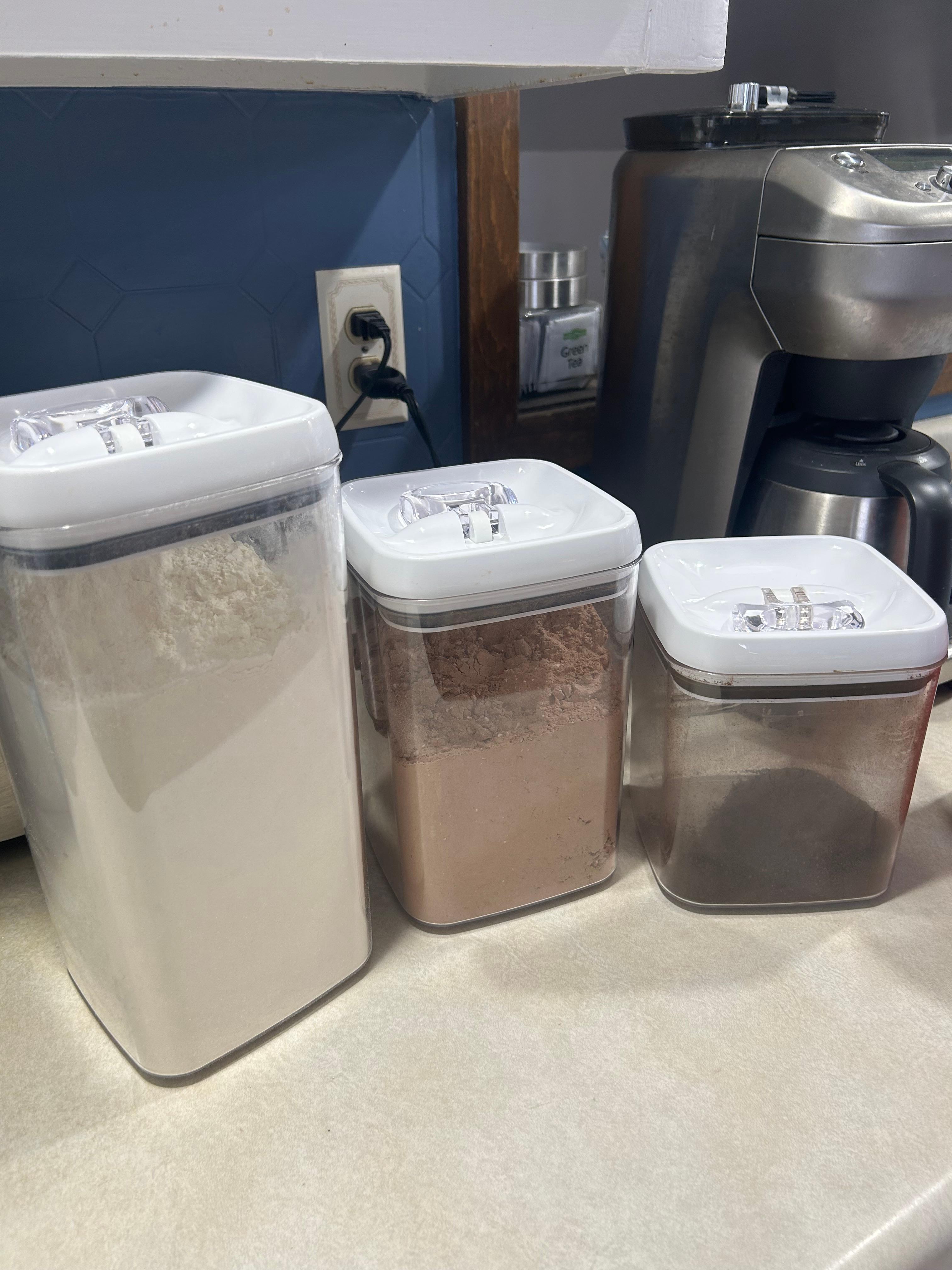 Several Better Homes & Gardens Flip-Tite containers on a kitchen counter, some empty and some filled with dry ingredients like flour and cocoa powder.