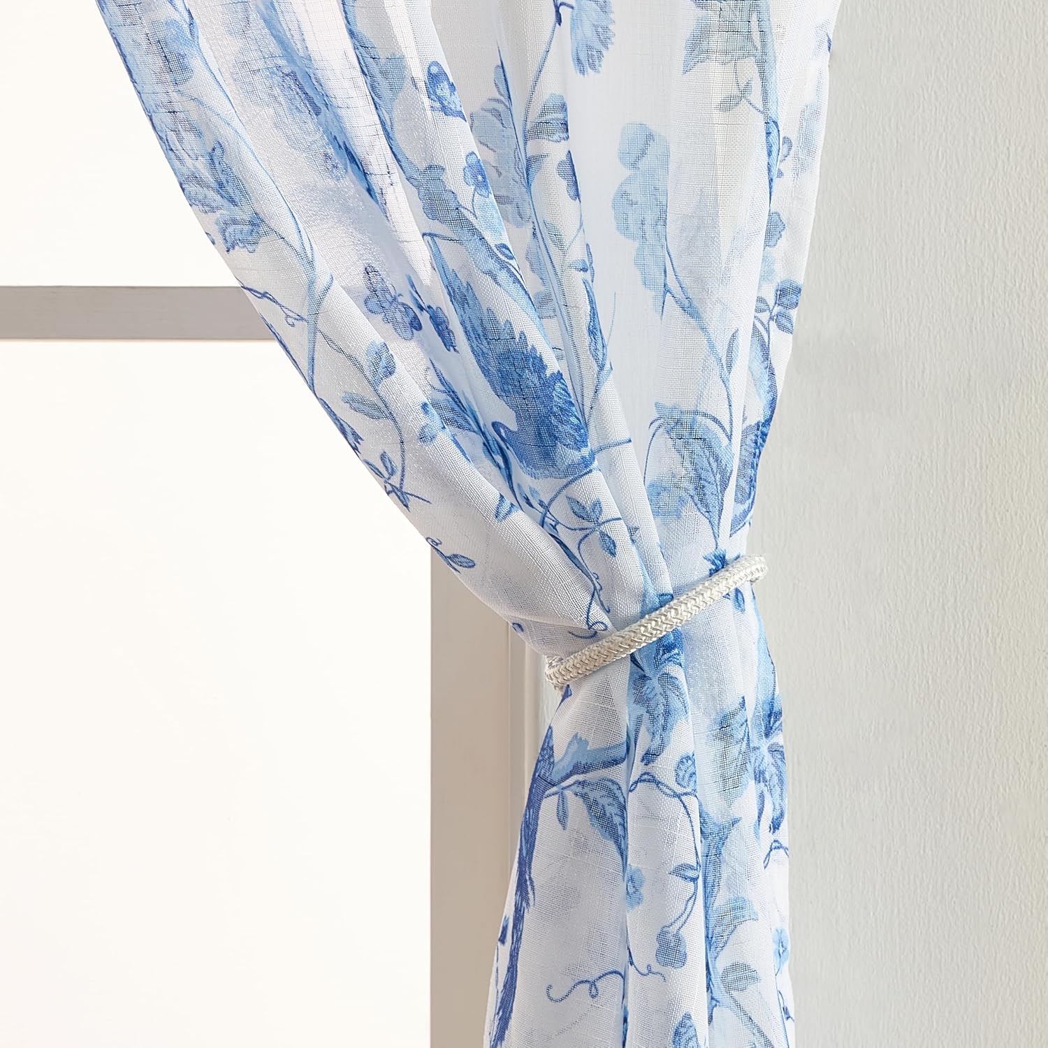 A sheer blue and white floral curtain panel tied back with a decorative silver tie-back.