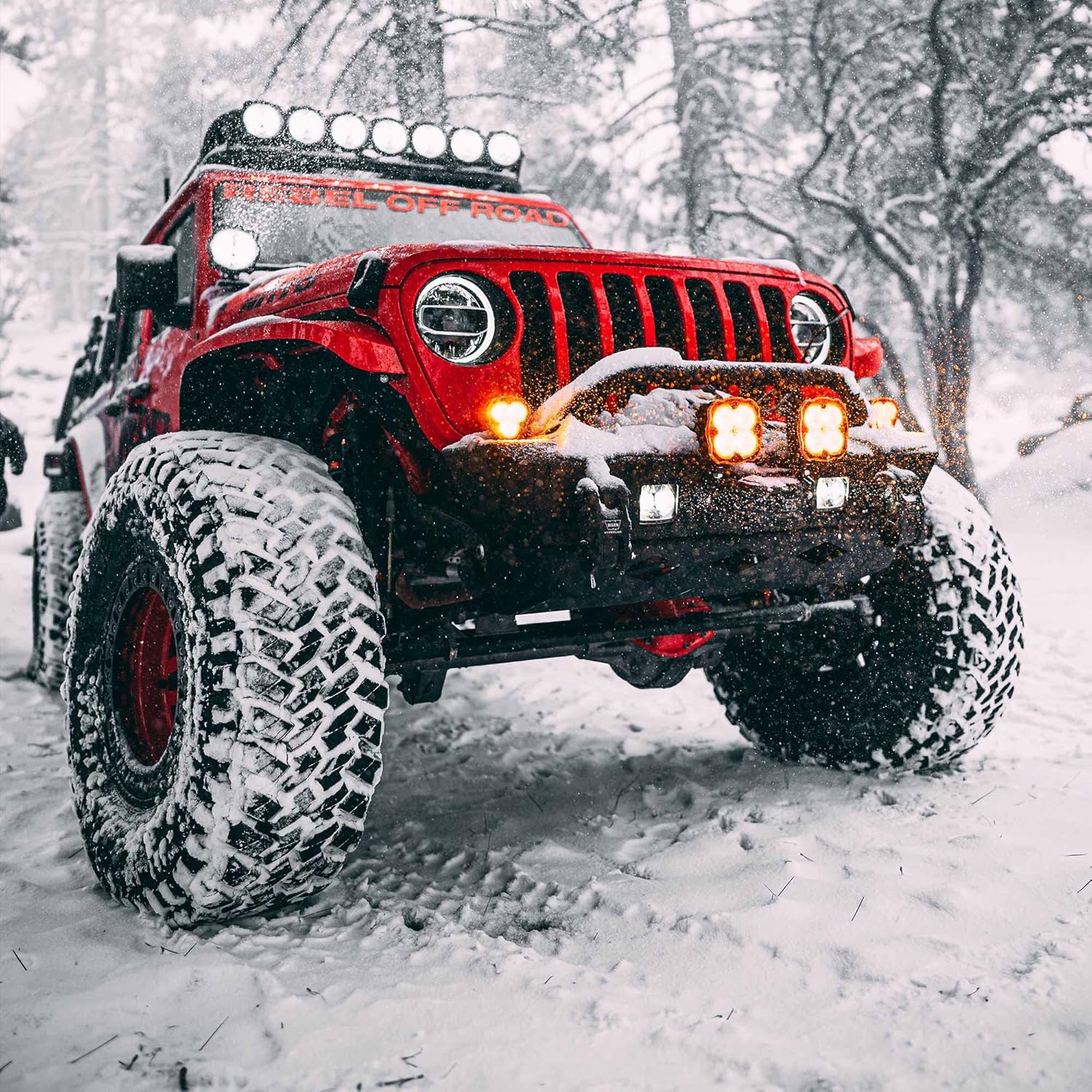 Red Jeep Wrangler equipped with multiple KC Hilites Flex ERA 4 LED light pods on the bumper and roof, driving through a snowy forest.