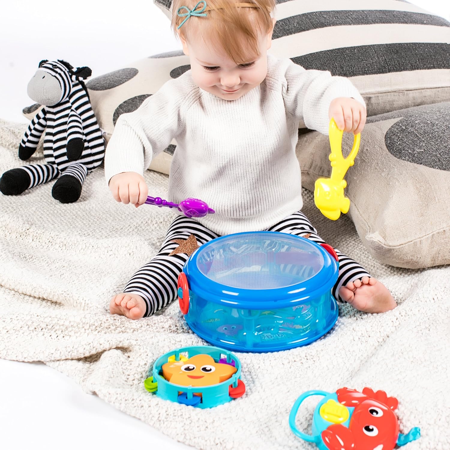 A baby sitting on a white blanket with striped pants, playing with the yellow fish maraca and the blue drum. A stuffed zebra toy is visible in the background.
