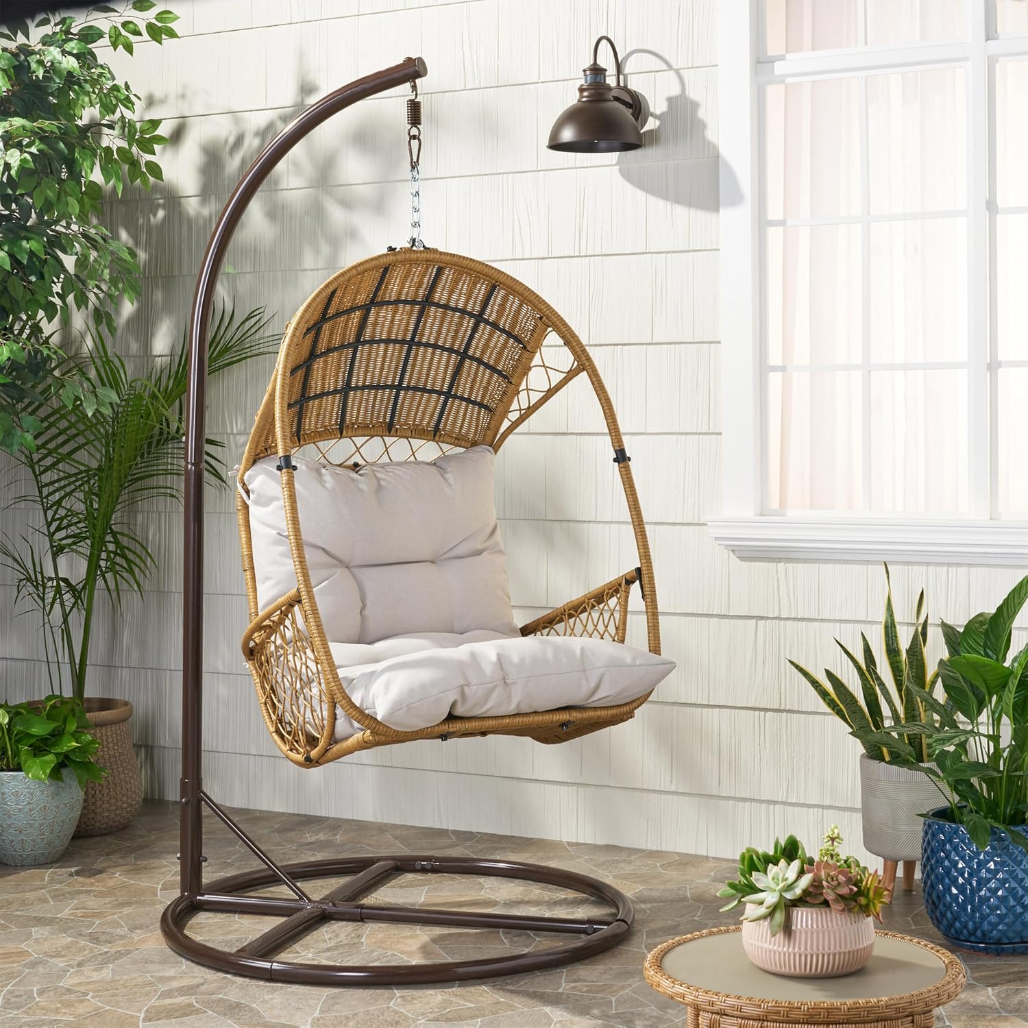 A GDFStudio Outdoor Wicker Hanging Chair with Stand, featuring a light brown wicker basket and a beige cushion, placed on a patio with potted plants.