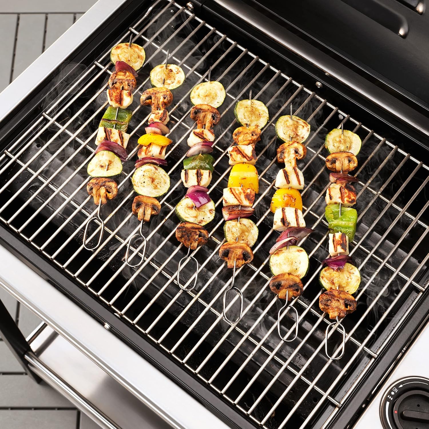 Several IKEA stainless steel grill skewers loaded with vegetables and cheese cooking on a barbecue grill.