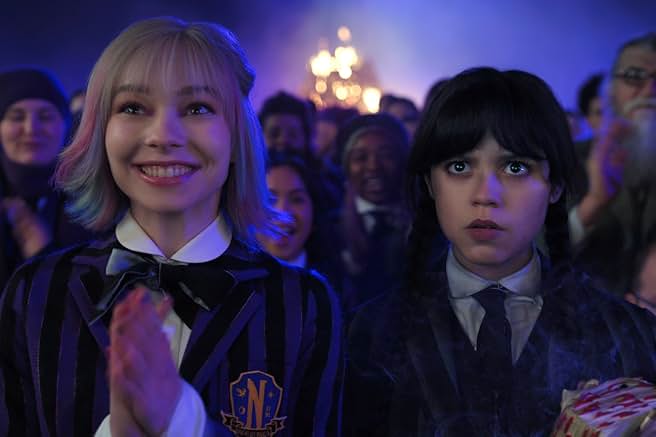 Jenna Ortega's Wednesday Addams and Emma Myers' Enid Sinclair watch Steve Buscemi's Barry Dort give a speech in front of a burning spire in the Here We Woe Again game