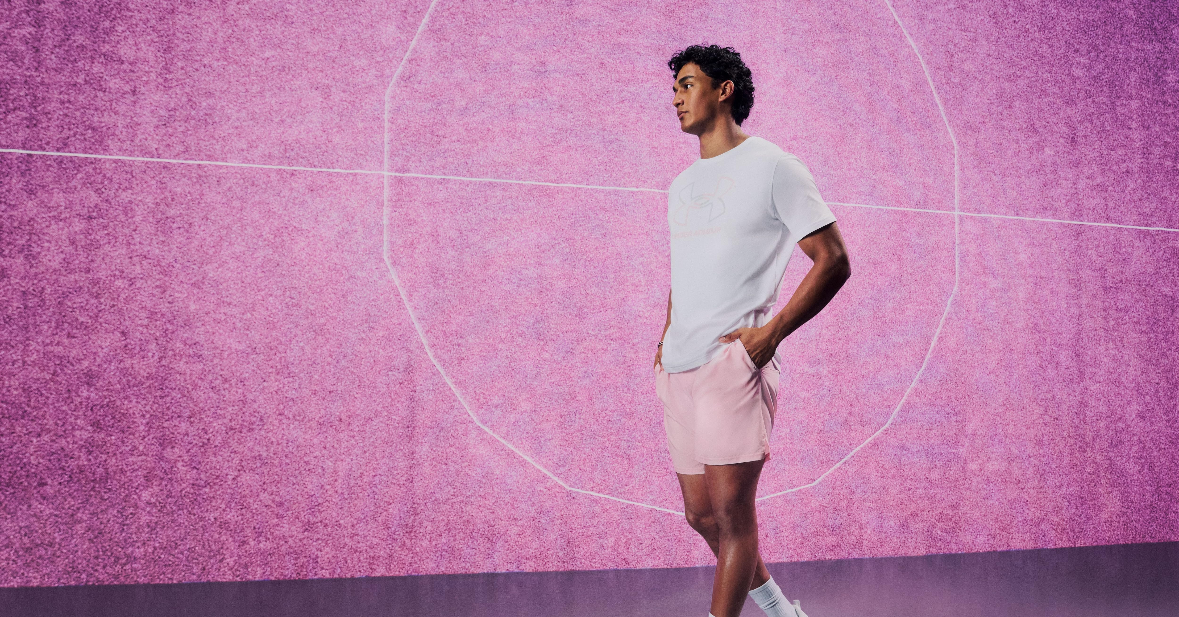 El texto dice «Imagen 1". Hombre vestido con camiseta blanca, pantalones cortos rosas y zapatillas blancas de pie sobre un fondo texturizado rosa con un patrón circular. Piso morado.