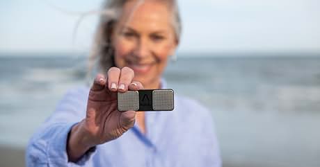 Pequeño dispositivo rectangular sostenido por una persona en la playa. El dispositivo parece ser una grabadora de audio portátil o un dispositivo electrónico similar.