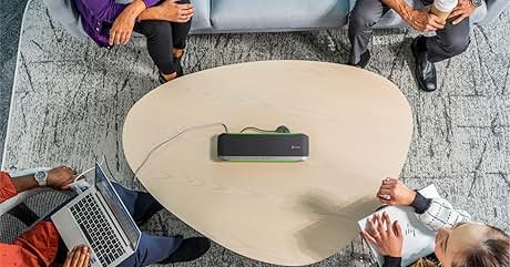 Vista aérea de una mesa de café curva de color beige con un dispositivo electrónico negro en el centro. Cuatro personas se sientan alrededor de ella sobre una alfombra gris estampada, y una usa una computadora portátil