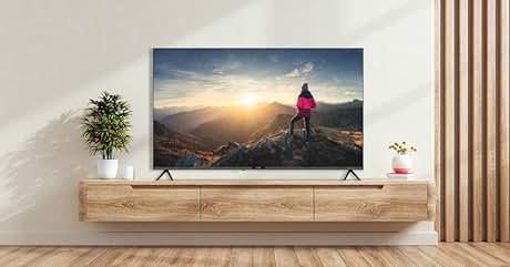 Gran televisor de pantalla plana montado en una pared blanca sobre una consola multimedia flotante de madera, que muestra una escena de puesta de sol en la montaña con una persona vestida con una chaqueta rosa, sala de estar moderna con plantas y cortinas.