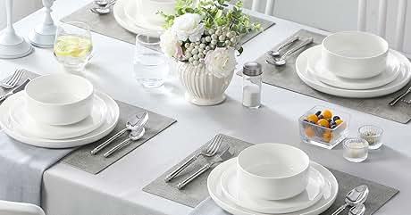 Elegant dining table set with white dishes, silver cutlery, and gray placemats. Centerpiece features white flowers in a vase. White chairs visible.