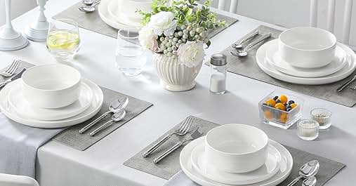 Elegant dining table set with white dishes, silver cutlery, and gray placemats. Centerpiece features white flowers in a vase. White chairs visible.