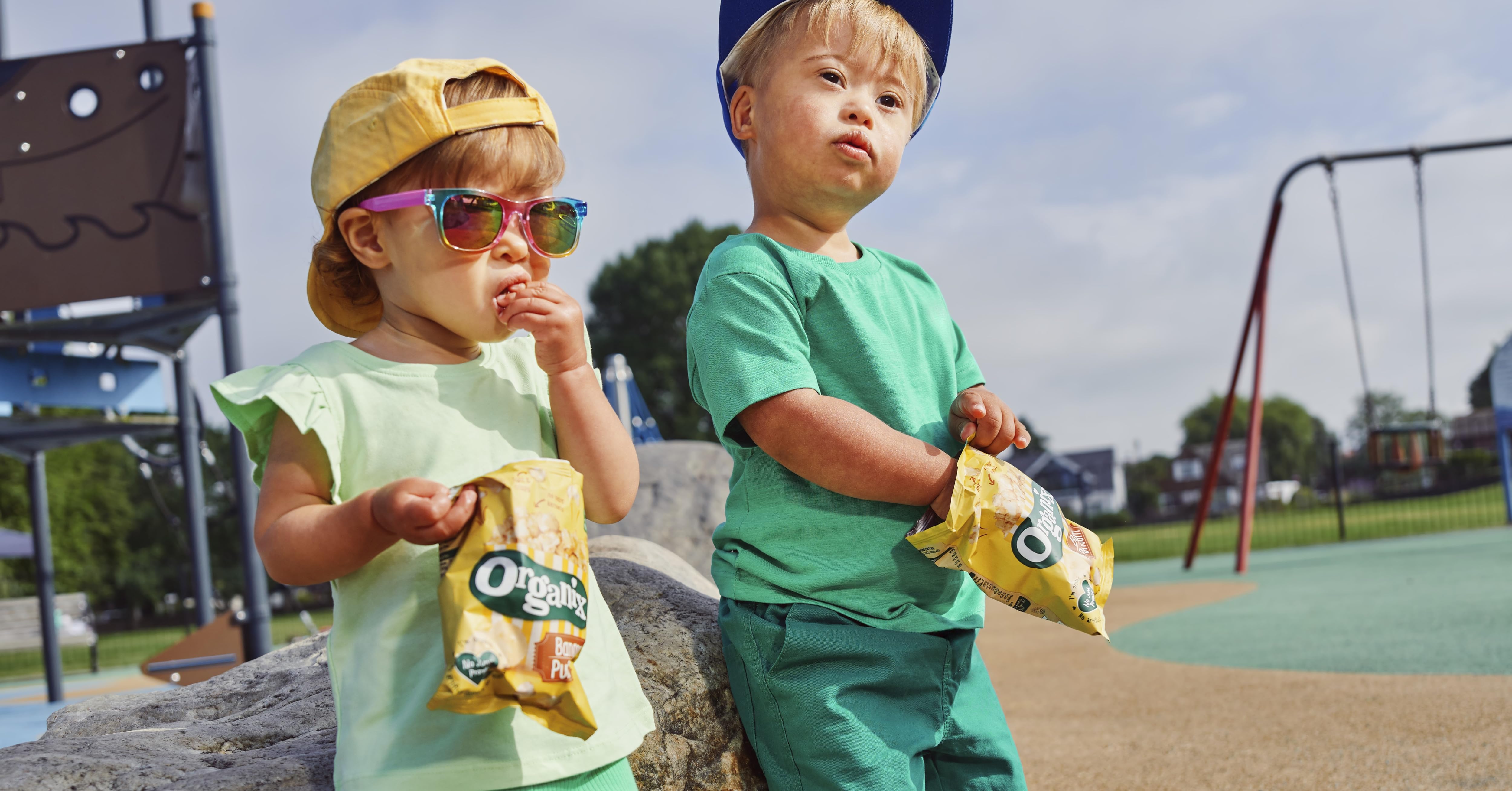 Two children outdoors eating yellow popsicles. One child wears colorful sunglasses.