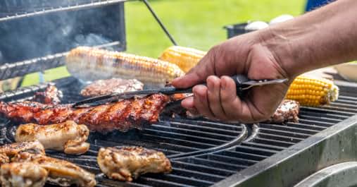 Barbecue grill with various meats cooking, including ribs and chicken. Hand holding tongs flips meat. Corn on the cob also grilling. Smoke rises from the grill.