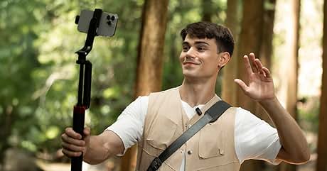Smartphone su selfie stick tenuto da una persona all'aperto. La persona indossa una camicia bianca e un gilet beige, salutando la telecamera. Sfondo boscoso visibile.