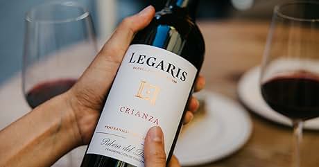 El texto dice «LEGARIS CRIANZA». Botella de vino con la etiqueta Legaris Crianza con la mano. Dos copas de vino con vino tinto visibles en el fondo sobre una superficie blanca