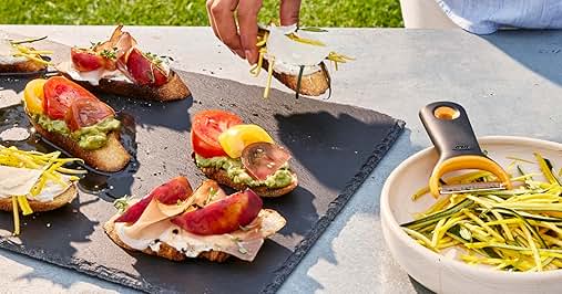 Open-faced sandwiches on dark slate serving board with colorful toppings including tomatoes and microgreens. Spiral vegetable garnish in white bowl alongside.