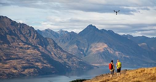 El texto dice «Imagen 1". Paisaje montañoso con lago. Dos personas vestidas con ropa colorida se paran en una colina cubierta de hierba con vistas al paisaje. Pequeño dron visible en el cielo. Picos escarpados y cielo nublado de fondo.