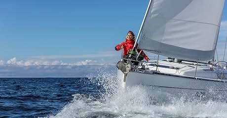 Velero surcando olas en aguas abiertas. La vela blanca ondea cuando el barco se inclina, con el rocío visible. Persona con chaqueta roja visible en cubierta