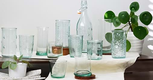 Collection of clear glass drinkware with various textures and patterns. Includes tumblers, bottles, and glasses in different sizes, arranged on a white surface with green plant accents.