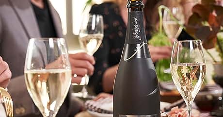 El texto dice «Freixenet». Botella de champán negro mate sobre la mesa del comedor con copas de vino, platos de comida y mesas