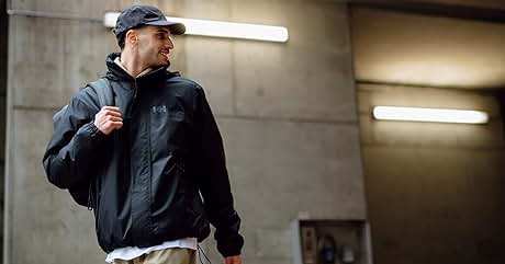 Hombre vestido con chaqueta negra con capucha y gorra de béisbol, de pie en un espacio interior de concreto con luces fluorescentes en lo alto.