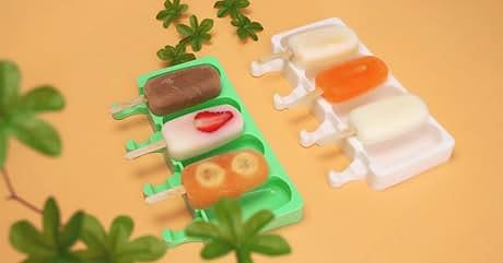 Assortment of colorful popsicle molds in green and white, featuring various shapes like fruit slices and classic popsicle forms. Displayed on an orange surface with decorative leaves.