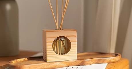 con base cuadrada y recorte circular, que muestra cañas delgadas. Colocado sobre una superficie de madera junto a la lámpara y el papel