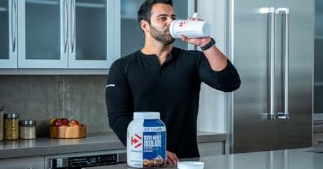 Man in black shirt drinking from a white shaker bottle in a kitchen. Holds a large container of Dymatize protein powder.