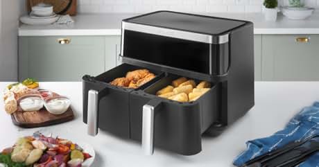 Black and silver dual-basket air fryer with digital display. Open drawers show fried chicken and bread rolls. Kitchen counter setting with food plates nearby.