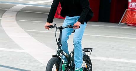 Una persona que conduce una bicicleta eléctrica plegable compacta con neumáticos gruesos. La bicicleta tiene un marco negro, detalles en verde y un portaequipajes trasero. El ciclista usa jeans y sostiene el manillar