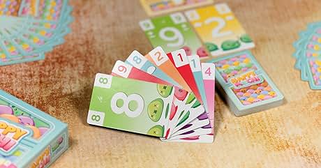 El texto dice «8", «1", «2", «3", «4". Se desplegaban coloridas tarjetas numeradas en forma de abanico, con ilustraciones de frutas. Cajas de cartón azules visibles en el fondo. Las tarjetas parecen ser parte de un juego infantil o juego educativo.