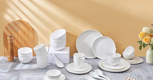 White ceramic dinnerware set including plates, bowls, cups, and utensils on gray surface. Wooden utensil holder, yellow flowers in vase, and beige backdrop with sunlight.