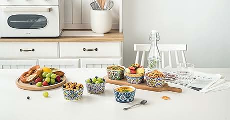 Kitchen counter scene with colorful patterned ceramic bowls in blue, yellow and geometric designs containing food, wooden cutting board with fruits and vegetables, white microwave, subway tile backsplash, and white dining chairs.